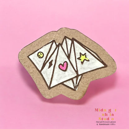 A wooden pin in the shape of a cootie catcher kid’s game in white with small bright symbols of a smile, lightning bolt, heart, and star