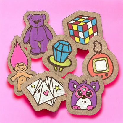 a collection of pins made of wood that are of 90's popular vintage retro toys.  the picture includes a purple beanie baby, a rubik's cube, a blue and lime green ring pop, and orange and yellow tamagotchia, a purple and pink furby, a cootie catcher, and a pink haired troll doll.  the background is pink.