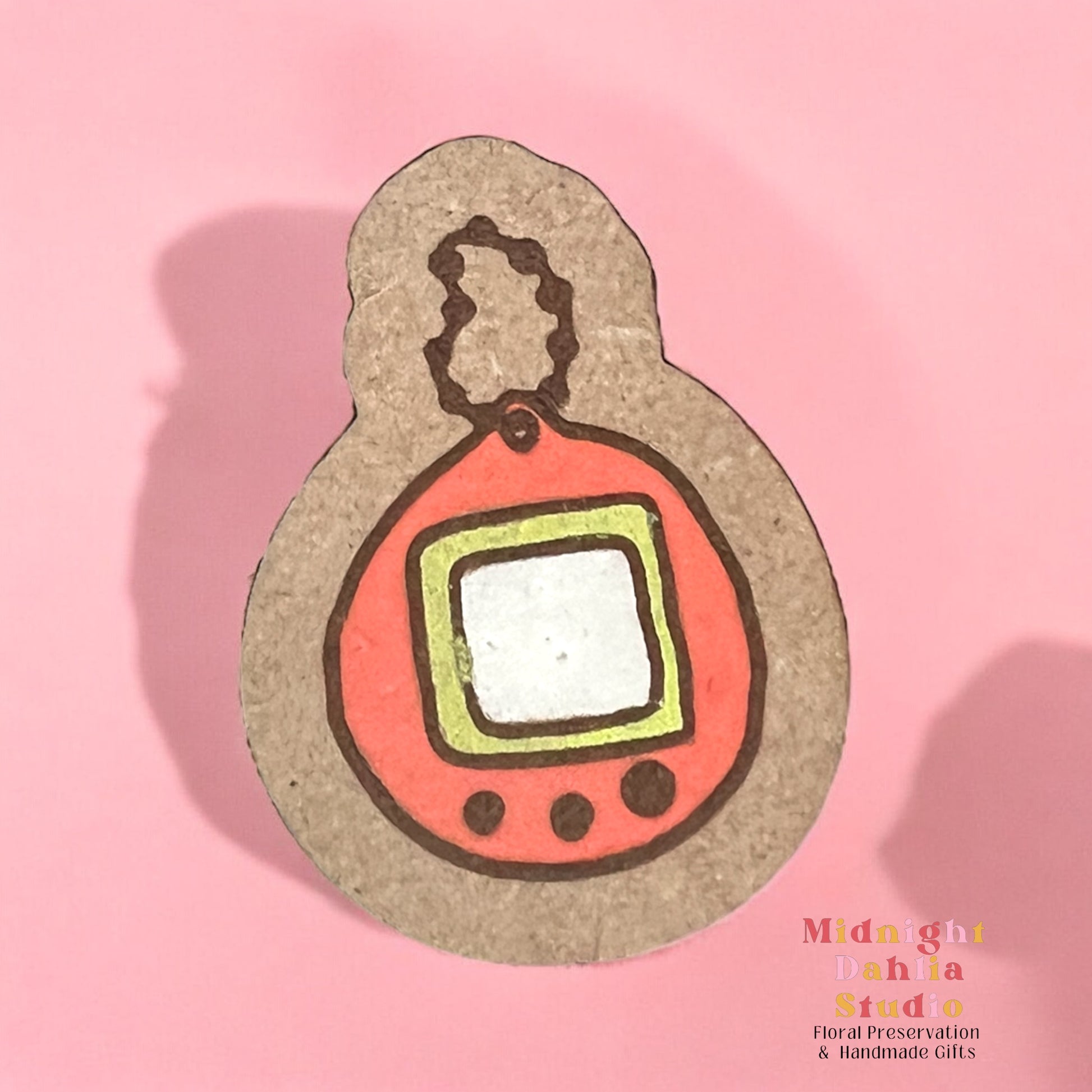 A wooden pin with a painted keychain in the style of a tamagotchi with an orange border and a small white screen surrounded by a yellow border with three buttons below the screen