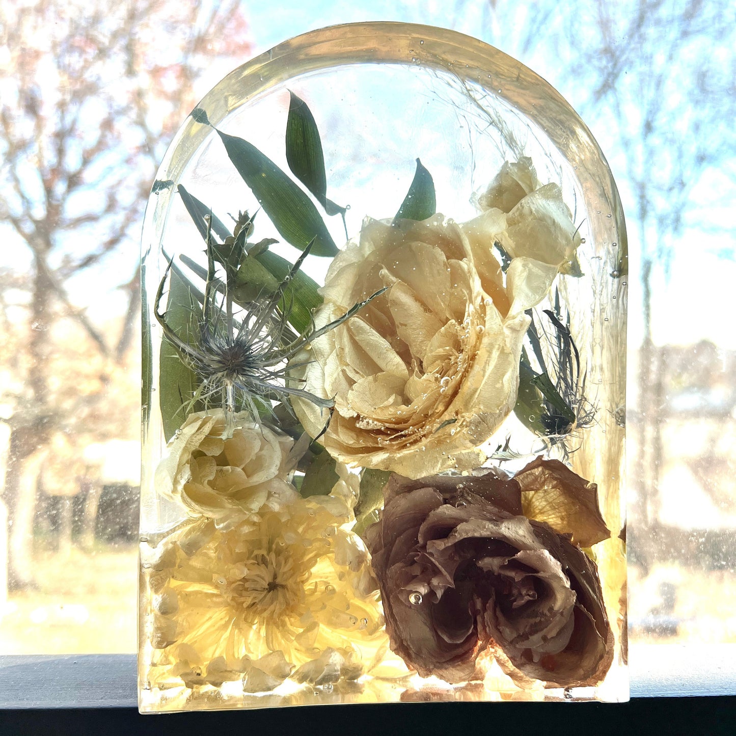 A custom crystal clear resin arch made with preserved flowers of brown taupe roses , white roses , white ranunculus , white mini spray roses , blue thistle , and green leaves .   Beautiful bouquet preservation option that helps you keep your bridal flowers forever.