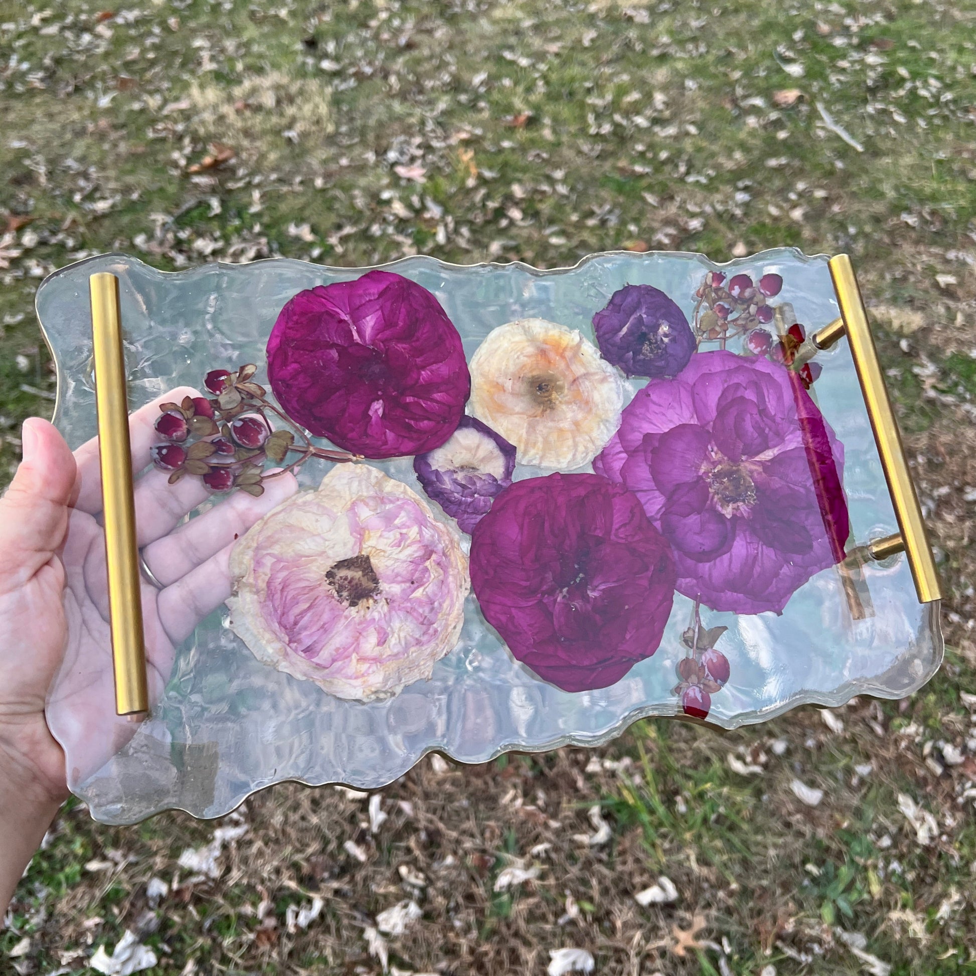 A crystal clear custom resin tray with gold handles with red and dark pink roses , red and purple ranunculus , and red hypericum berries.  This is a beautiful addition to a custom bouquet preservation order.