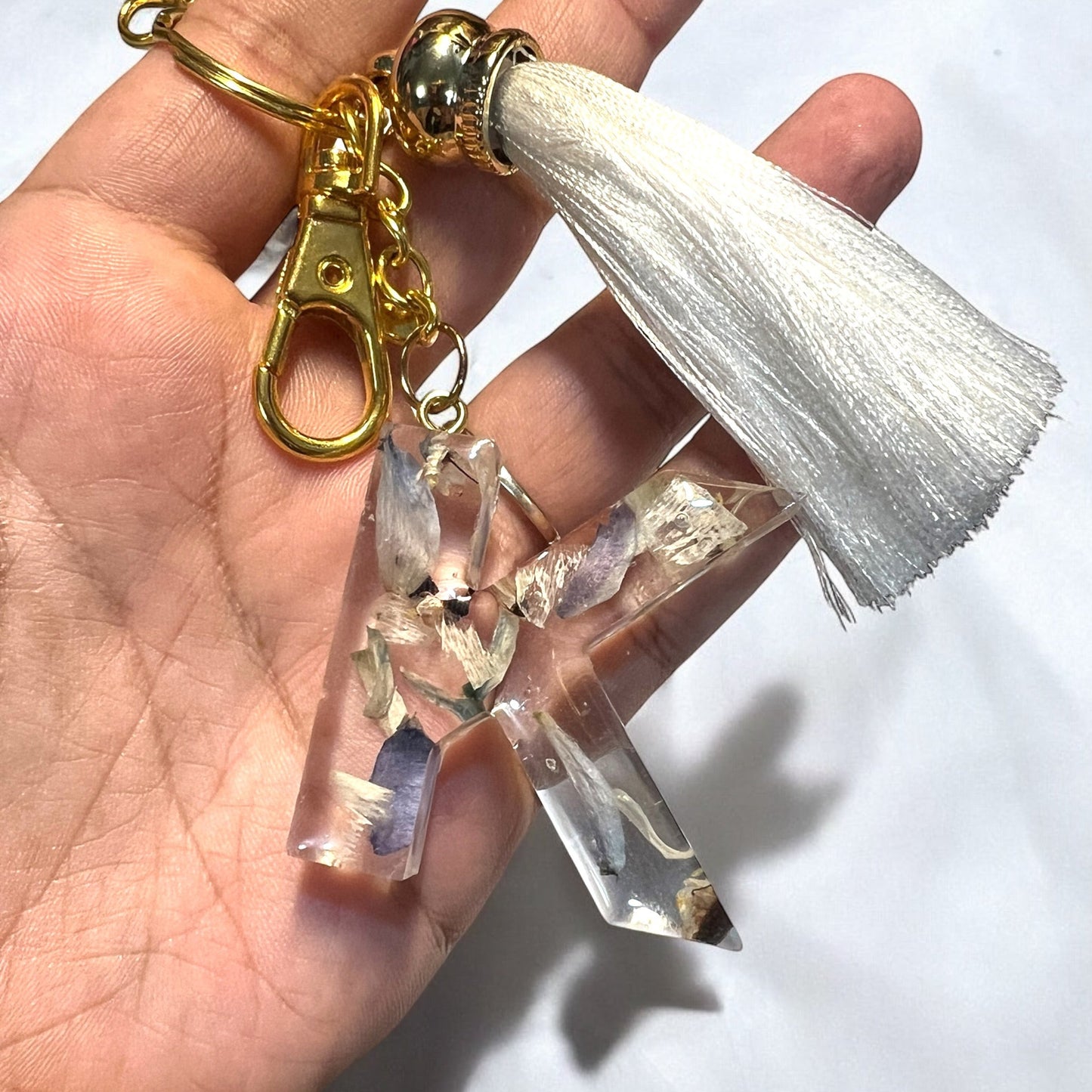 A custom resin letter keychain in the shape of a K with tiny blue and white flowers.  The keychain has gold hardware and an off-white tassel.  It is a beautiful addition to a bridal bouquet preservation.