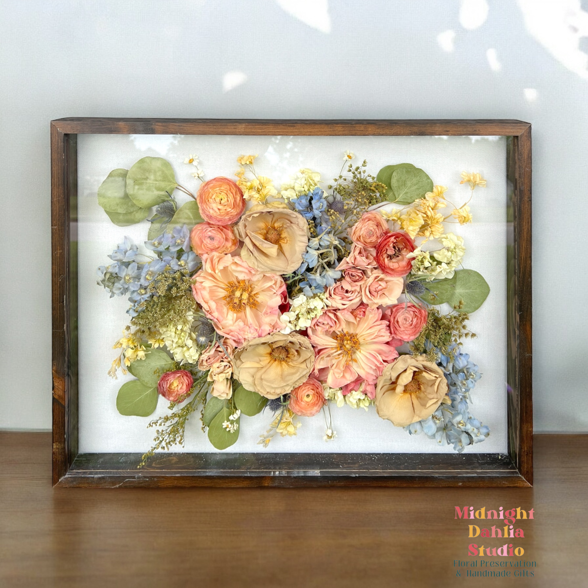 a wooden framed shadow box filled with many flowers, including peach peonies, taupe roses, white stock, blue delphinium, coral peach ranunculus, white stock, white hydrangea, yellow solidago, and silver dollar eucalyptus