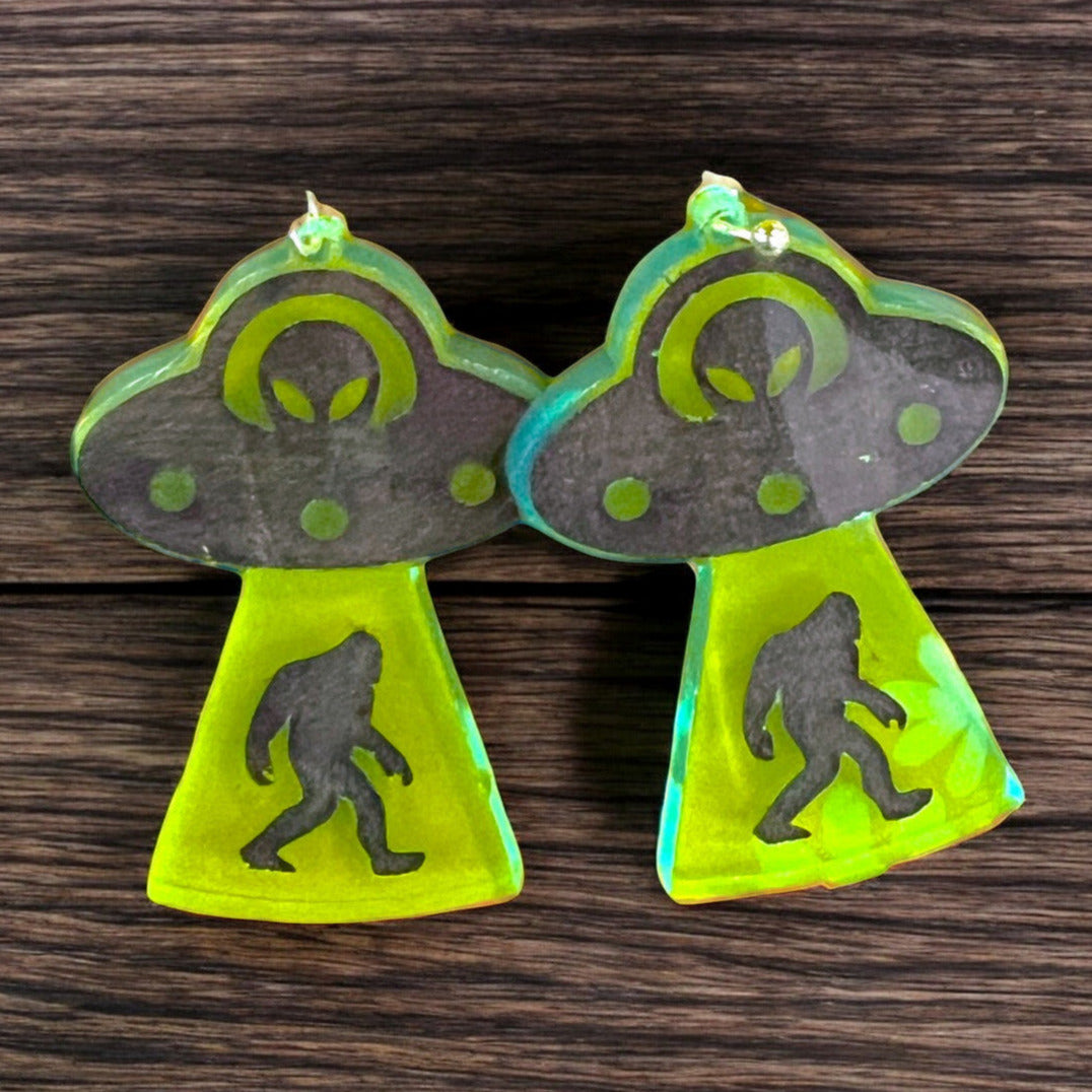 A pair of dangle earrings that depict a ufo with an alien driver abducting a bigfoot.  the earrings are made of neon green acrylic and are a perfect gift for a woman that loves cryptids and silliness.