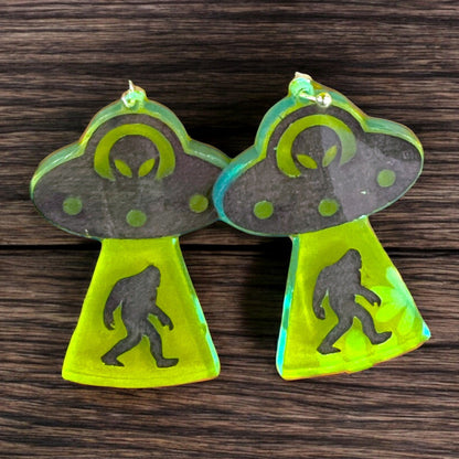 A pair of dangle earrings that depict a ufo with an alien driver abducting a bigfoot.  the earrings are made of neon green acrylic and are a perfect gift for a woman that loves cryptids and silliness.