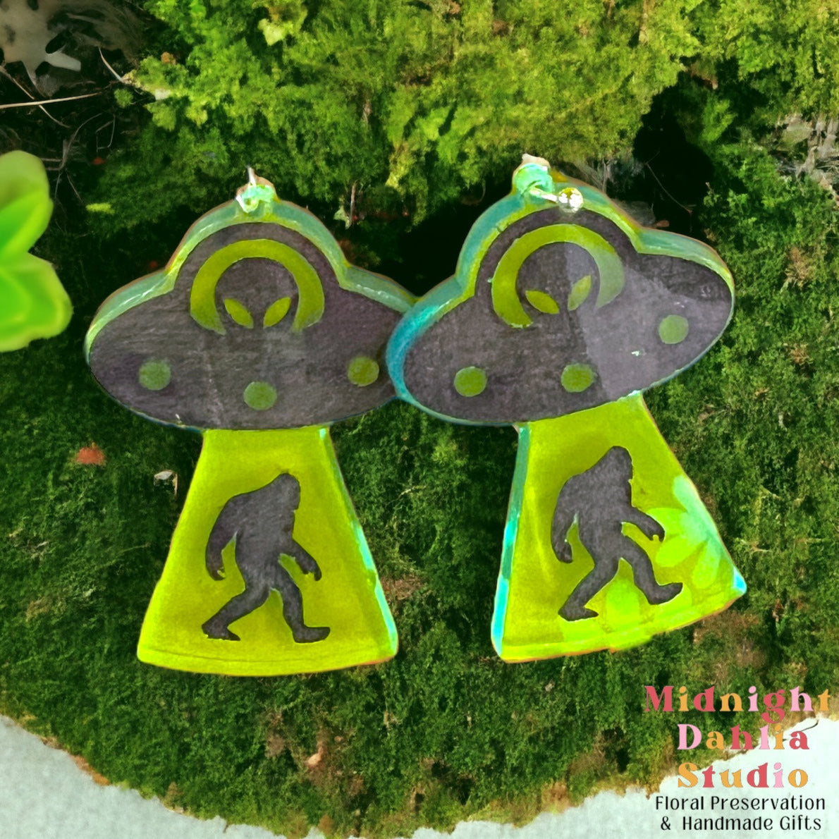 A pair of dangle earrings that depict a ufo with an alien driver abducting a bigfoot.  the earrings are made of neon green acrylic and are a perfect gift for a woman that loves cryptids and silliness.