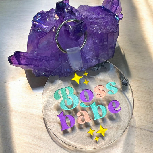 A round clear acrylic keychain that reads Boss Babe in teal green, purple, and peach, with yellow stars above and below the phrase.  