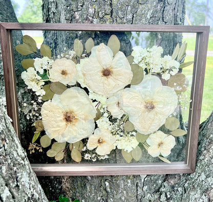 A custom pressed flower floating frame with preserved white roses , white hydrangea , and green eucalyptus .   A beautiful way to preserve a bridal bouquet.