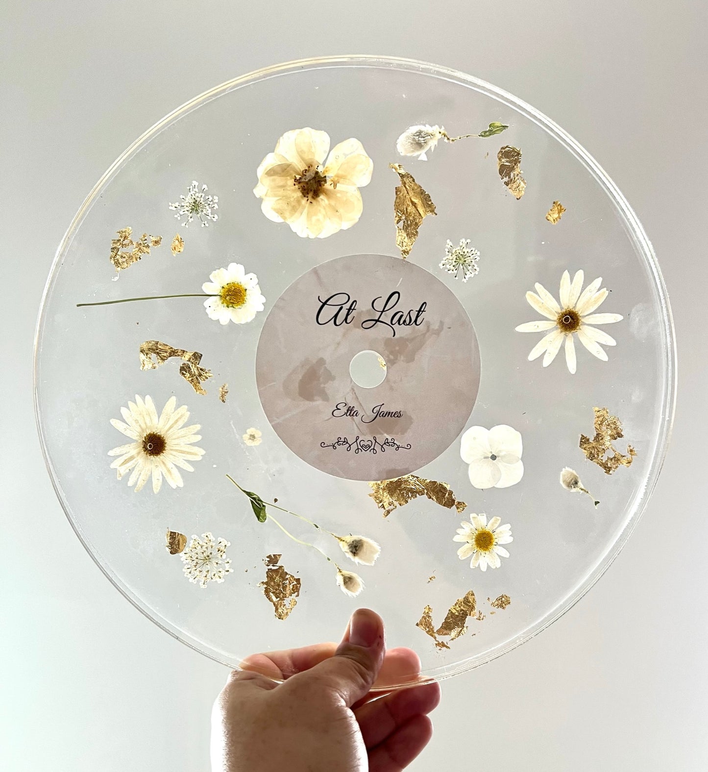 A custom decorative resin record made with white daisies , white roses , white hydrangea , and green leaves , and gold flake.  It has a label to make it look like an album.