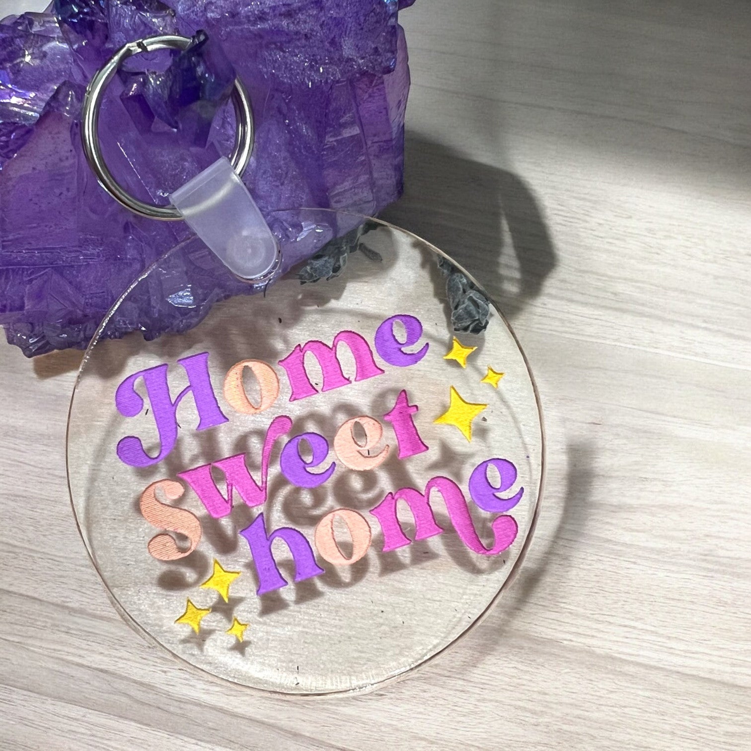 a round clear acrylic keychain with home sweet home etched on it in purple, peach orange, and pink with yellow stars on either side of the words.