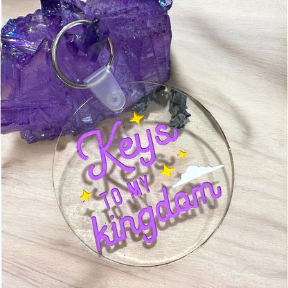 a round clear acrylic keychain that reads keys to my kingdom in purple with a small white cloud and yellow stars surrounding the words.