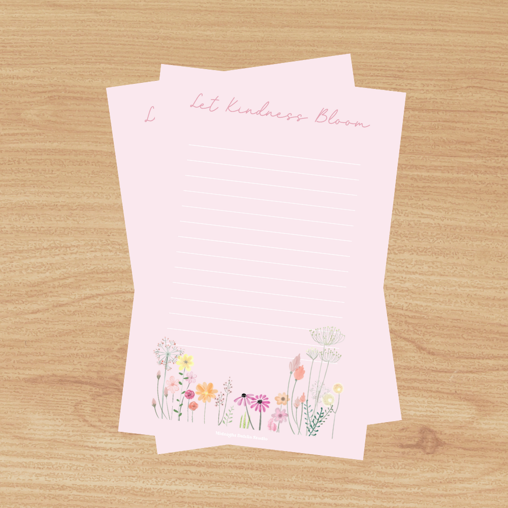 a pink lined notepad that reads let kindness bloom at the top in cursive script and wildflowers along the bottom