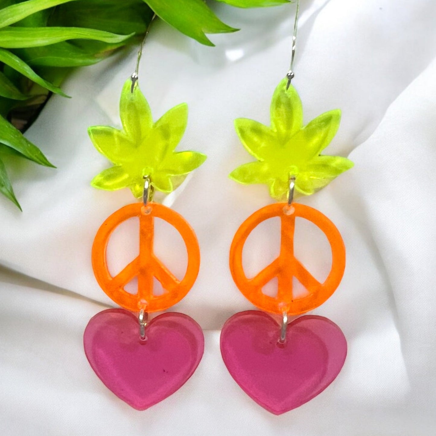 a trio of acrylic pieces hanging from silver plated earring hardware in the shape of a green marijuana pot leaf, an orange peace sign, and a pink heart.