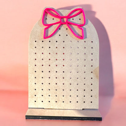 a jewelry organizer made of wood with holds laser cut and a hot pink acrylic bow at the top