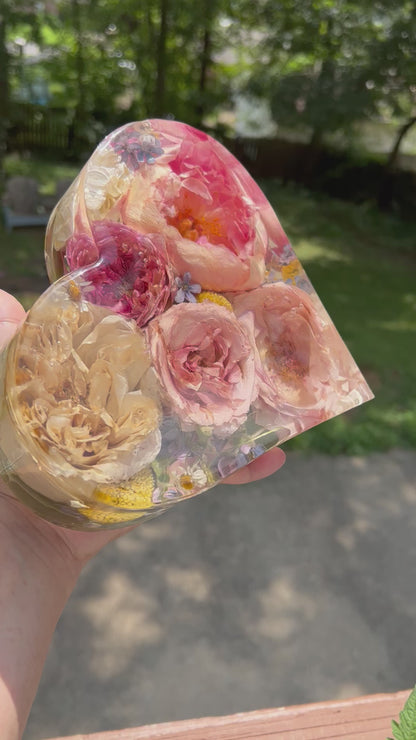 A crystal clear resin heart filled with pink and white roses , a pink peony , and other yellow and blue flowers . A beautiful way to preserve a bridal bouquet.