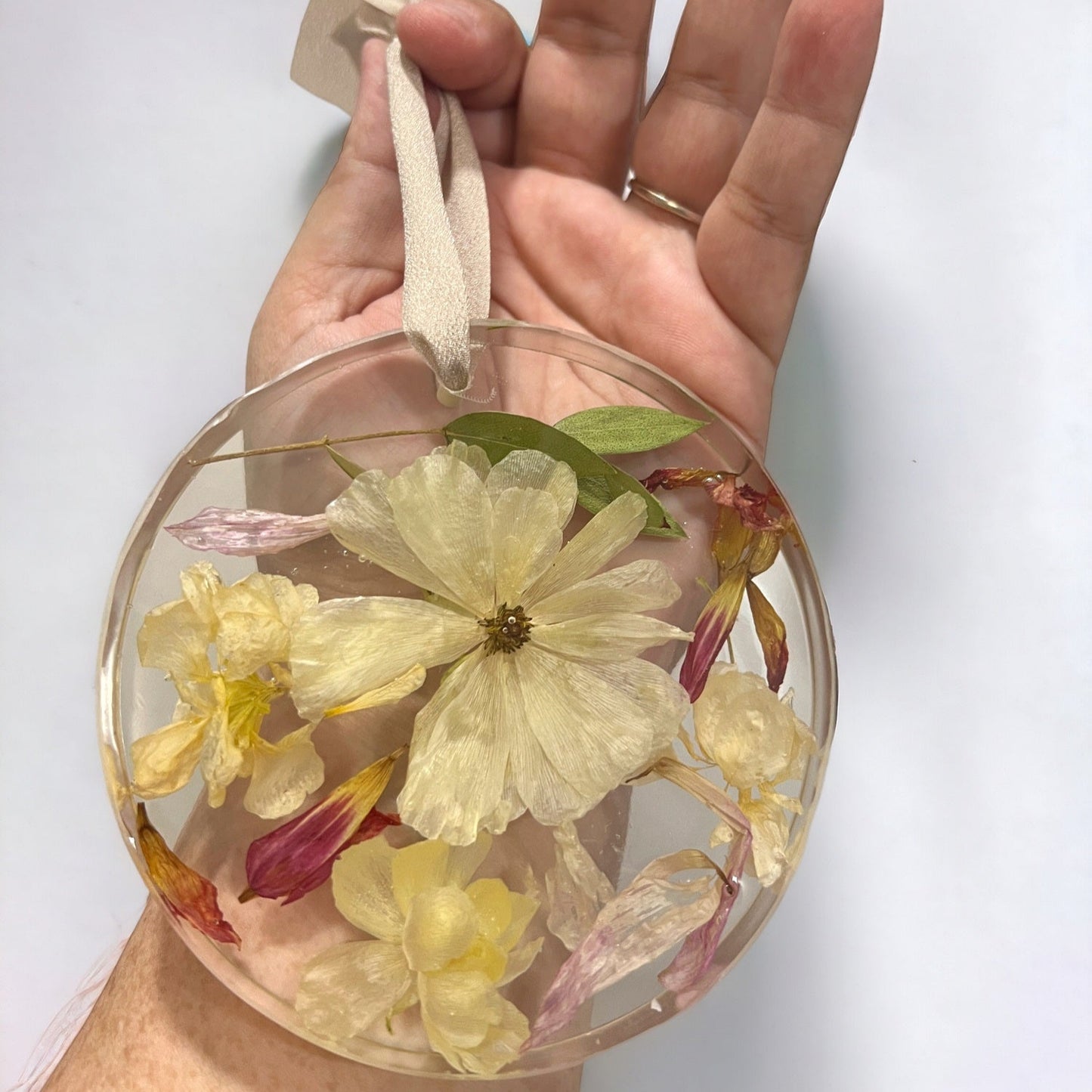 A crystal clear circle resin christmas ornament with white and yellow ranunculus , pink petals , and green leaves , with an off-white ribbon.  A beautiful add-on for a bridal bouquet preservation.