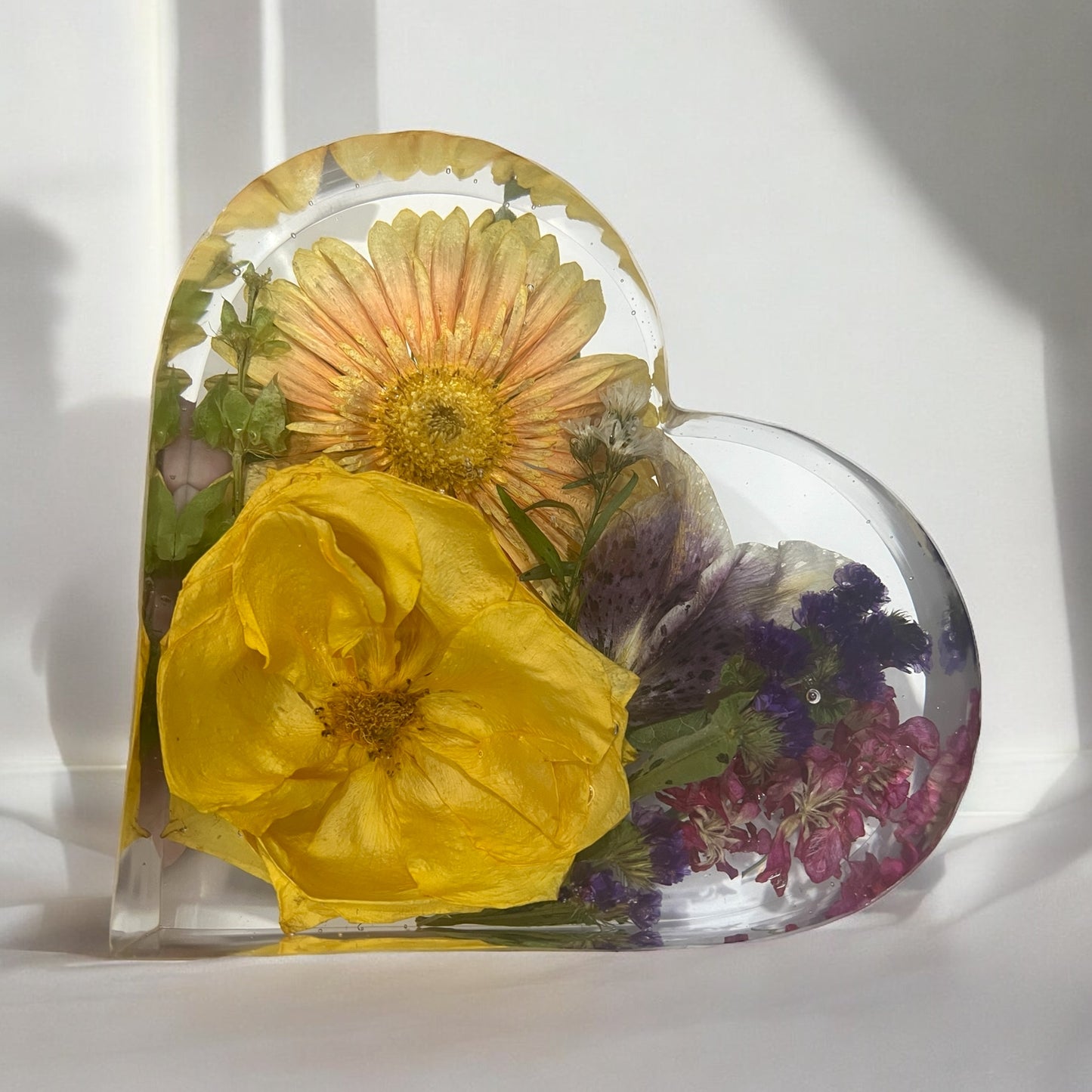 A crystal clear resin heart filled with a yellow rose , an orange gerber daisy , a lily , and other purple flowers . A beautiful way to preserve a bridal bouquet.