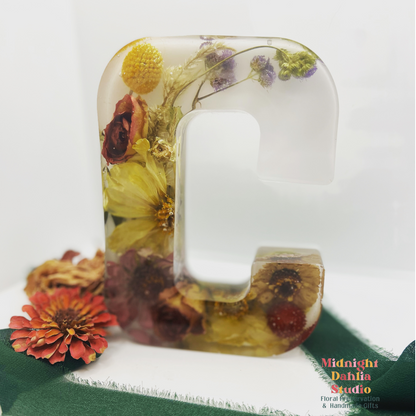 A crystal clear resin letter C with dried preserved flowers , including yellow billy balls , red roses , and a yellow dahlia .  A beautiful way to preserve your bridal bouquet.