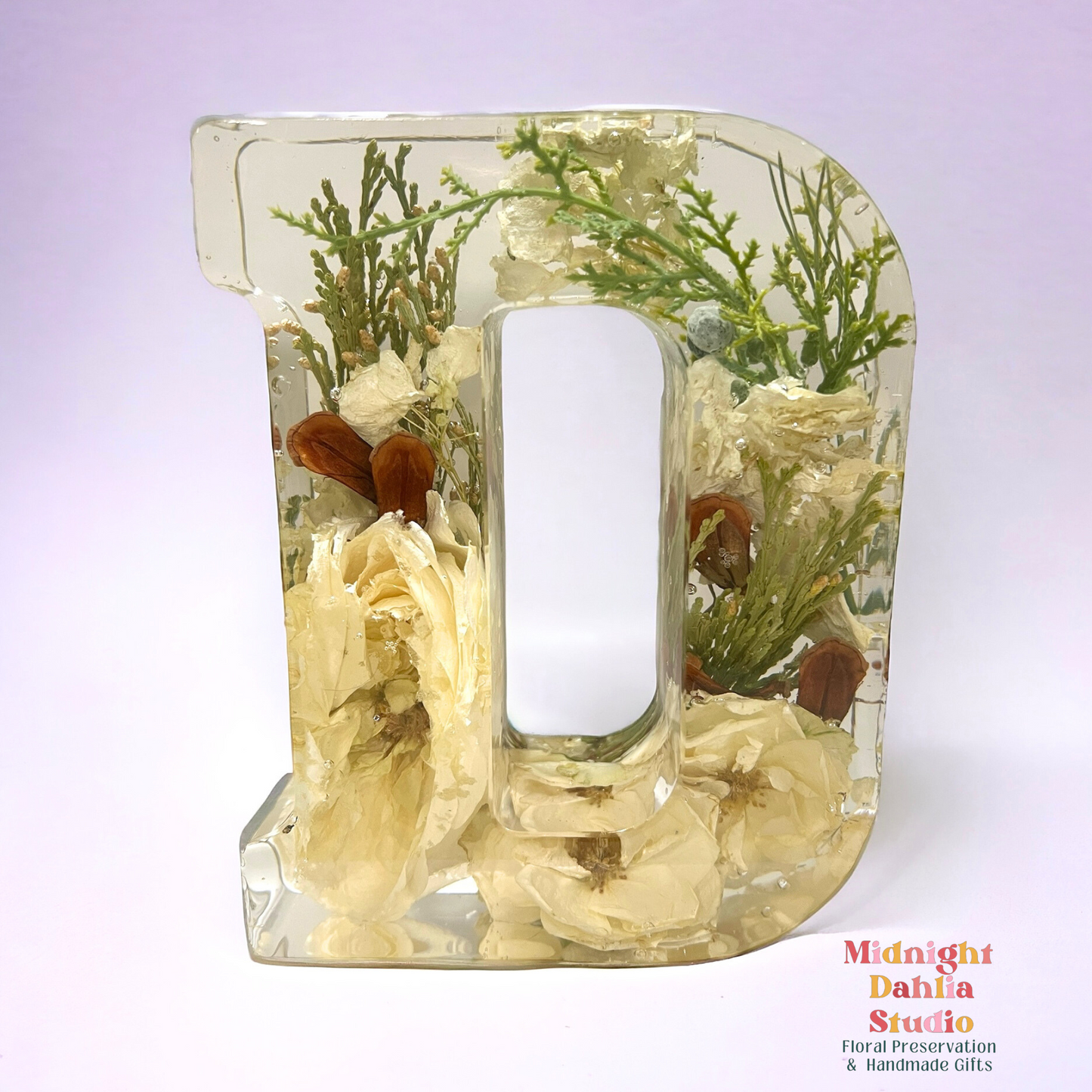 A crystal clear resin letter D with dried preserved flowers , including white roses , cedar , and pine cone pieces .  A beautiful way to preserve your bridal bouquet.