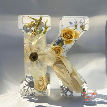 A crystal clear resin letter K with dried preserved flowers , including a white anemone , white and yellow roses , and blue delphinium and tweedia .  A beautiful way to preserve your bridal bouquet.