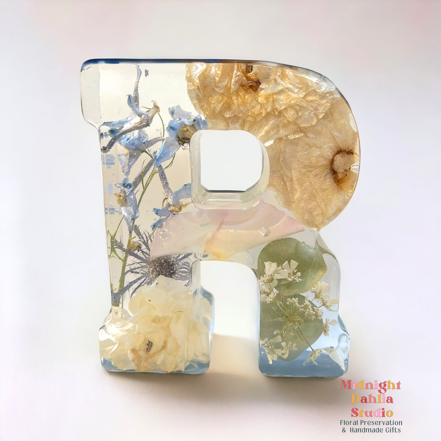 A crystal clear resin letter with dried preserved flowers , including a rose , a ranunculus , blue delphinium , blue thistle , babys breath , and green leaves.  A beautiful way to preserve your bridal bouquet.