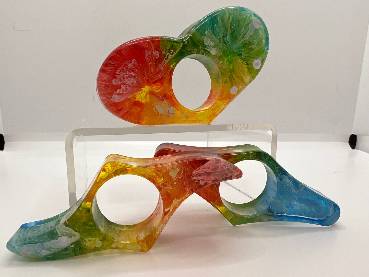 three book page holders on a white background. one is the shape of a heart sitting on a clear acrylic riser and there are two in front of the riser. each book page holder is rainbow in red, orange, yellow, green, and blue with white swirls.